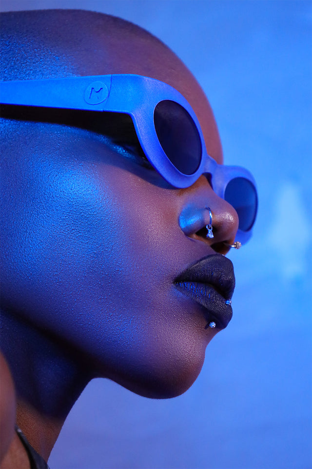 Female model in bold blue lighting wearing white oval REFRAMD sunglasses