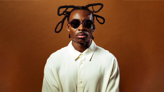 Stylish portrait of a man with unique braids and round sunglasses, exuding modern fashion against a warm background.