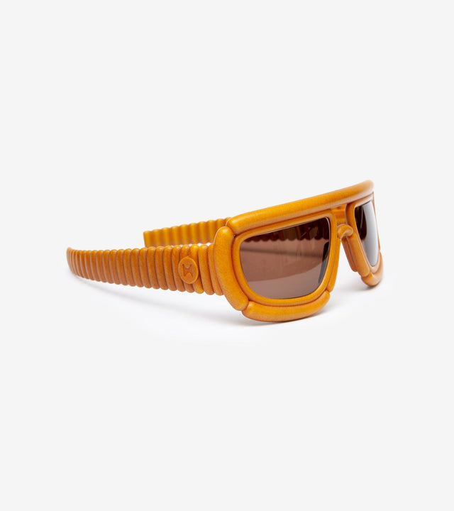 Side view of unique orange coiled-arm sunglasses with brown tinted lenses against a white background, highlighting the innovative design and vibrant color