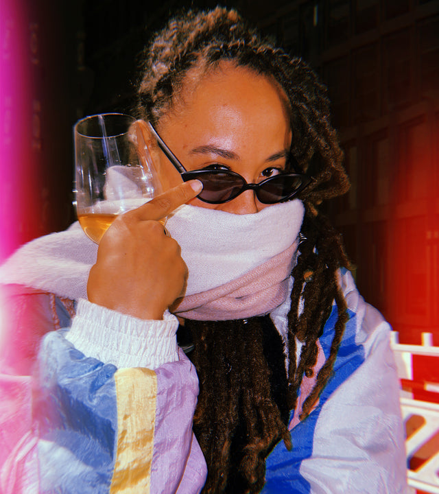 Cozy indoor scene with a person sipping beer, wearing narrow black sunglasses and a white scarf over their mouth, highlighted by dreadlocks and a color-blocked jacket against a warm-toned backdrop