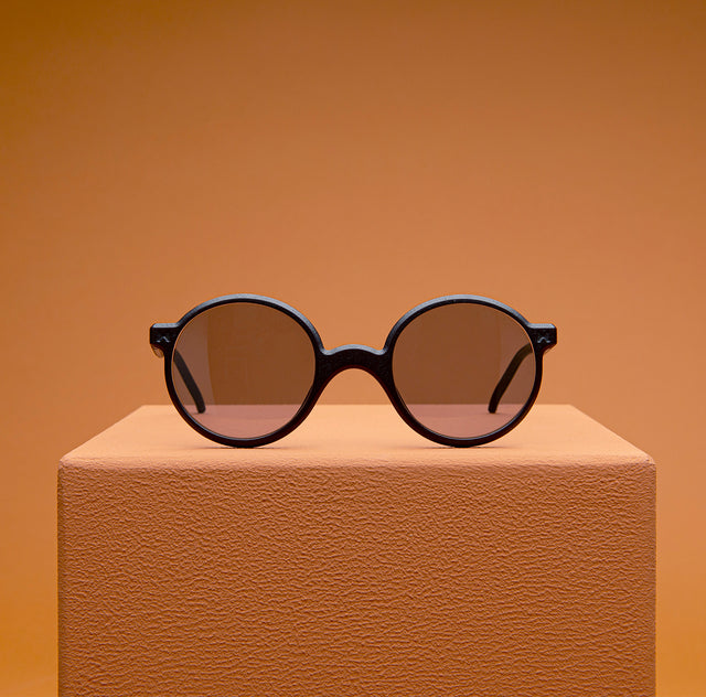 Round, black-framed sunglasses is elegantly displayed on an orange pedestal - sophisticated and modern aesthetic