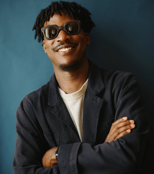 Portrait of a smiling man with stylish sunglasses and short dreadlocks, dressed in a navy worker blazer and cream sweater, set against a serene blue background.